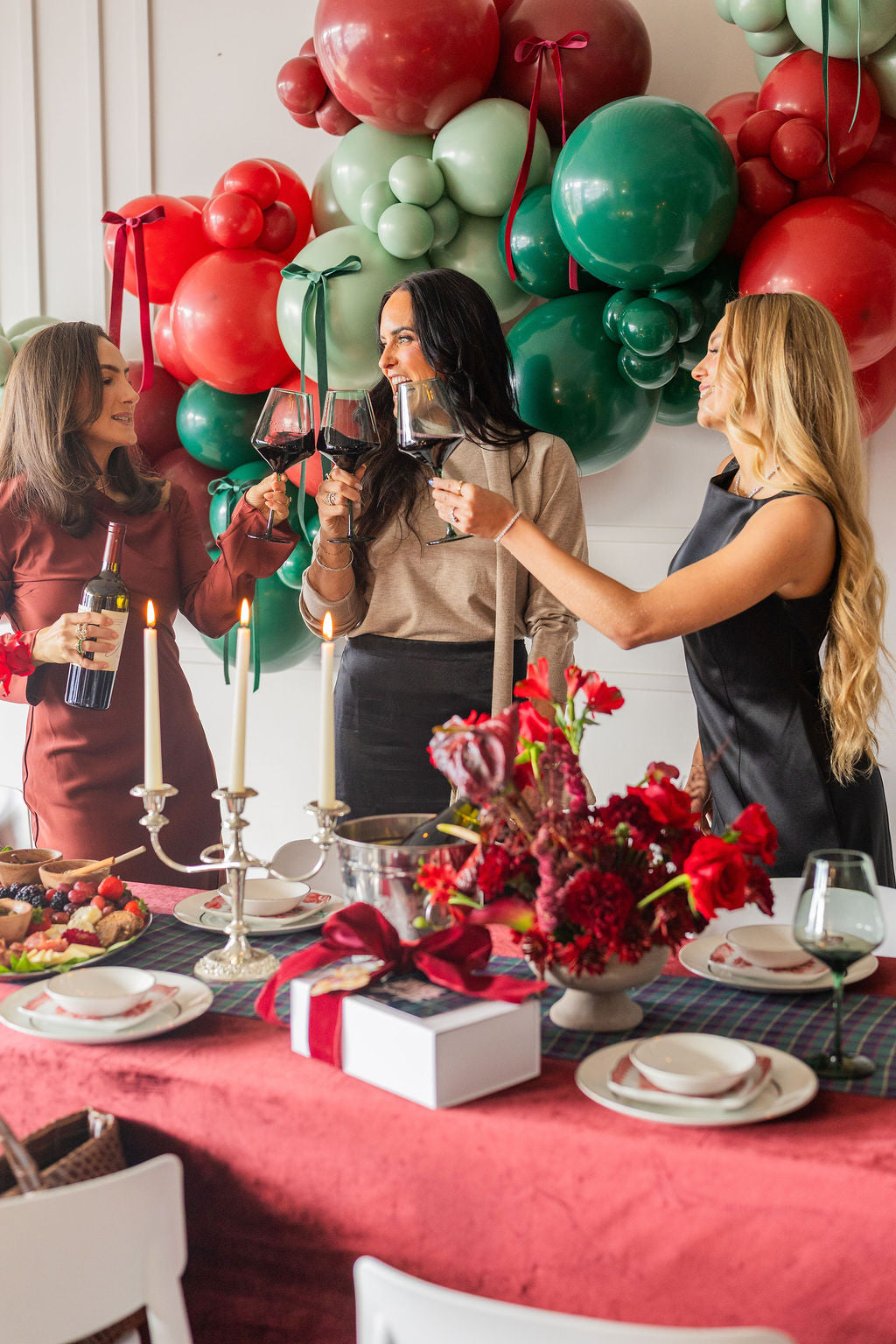 Three women celebrating with wine beside festive red and green balloons and a holiday table